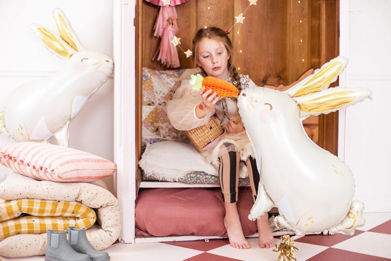 May include: A young girl sits in a white wooden cubby with a white and gold bunny balloon. She is holding a carrot-shaped balloon and a wicker basket. The cubby is decorated with pink and white pillows and blankets.