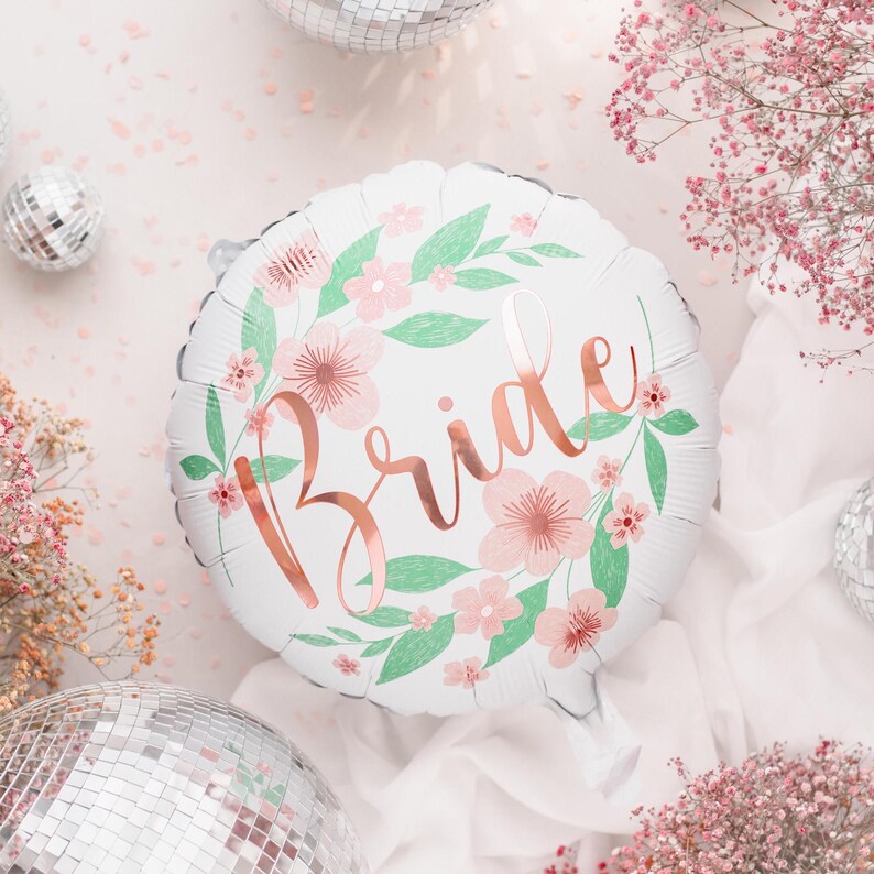 May include: A white round balloon with the word "Bride" in rose gold script. The balloon is decorated with a floral wreath of pink flowers and green leaves. The background includes disco balls and pink flowers.