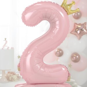 May include: Pink number two balloon with a gold crown on top. The balloon is standing upright on a white surface.