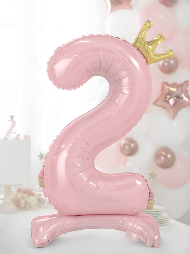 May include: Pink number two balloon with a gold crown on top. The balloon is standing upright on a white surface.
