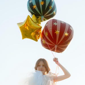 May include: A young person holds three balloons against a bright sky. One is a gold star, another is red with white stripes and stars, and the third is teal with black stripes and stars. The balloons are festive and colorful.
