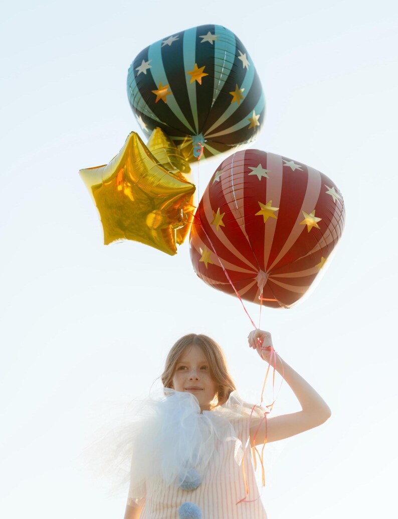 May include: A young person holds three balloons against a bright sky. One is a gold star, another is red with white stripes and stars, and the third is teal with black stripes and stars. The balloons are festive and colorful.