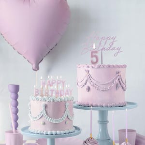 May include: Two pink frosted cakes with pink candles on top. The cakes are decorated with white frosting and have a pink heart balloon in the background. The text "Happy Birthday" is on the candles.