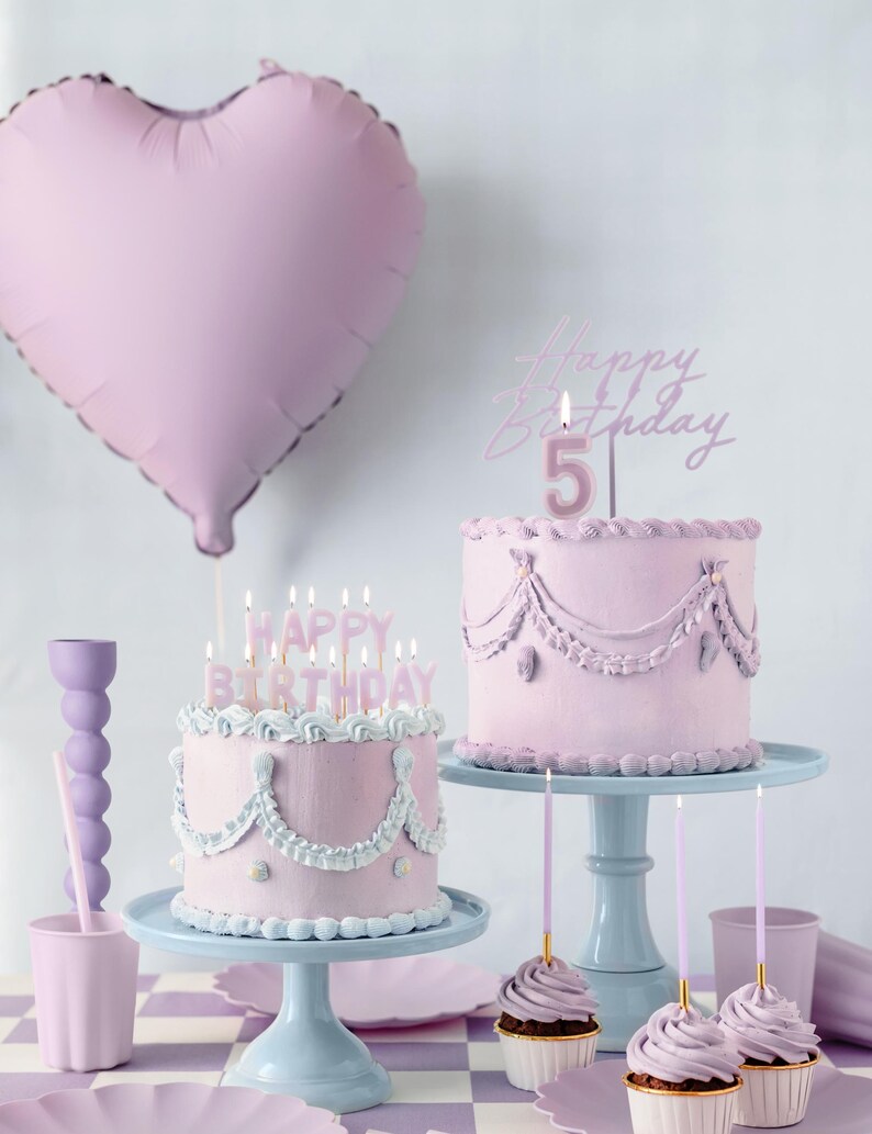May include: Two pink frosted cakes with pink candles on top. The cakes are decorated with white frosting and have a pink heart balloon in the background. The text "Happy Birthday" is on the candles.