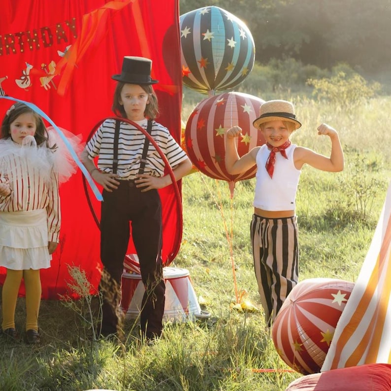 K&ouml;nnte beinhalten: Drei Kinder in Zirkuskost&uuml;men posieren im Freien. Eines tr&auml;gt einen Zylinder und ein gestreiftes Hemd, ein anderes ein wei&szlig;es Oberteil und eine gestreifte Hose, und das dritte einen R&uuml;schenkragen. Ballons mit Sternenmustern sind im Hintergrund.