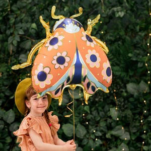 May include: A young person holds a large orange, blue, and gold ladybug balloon with floral patterns. The balloon is in front of a green leafy wall with string lights.