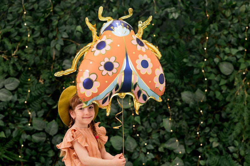 May include: A young person holds a large orange, blue, and gold ladybug balloon with floral patterns. The balloon is in front of a green leafy wall with string lights.