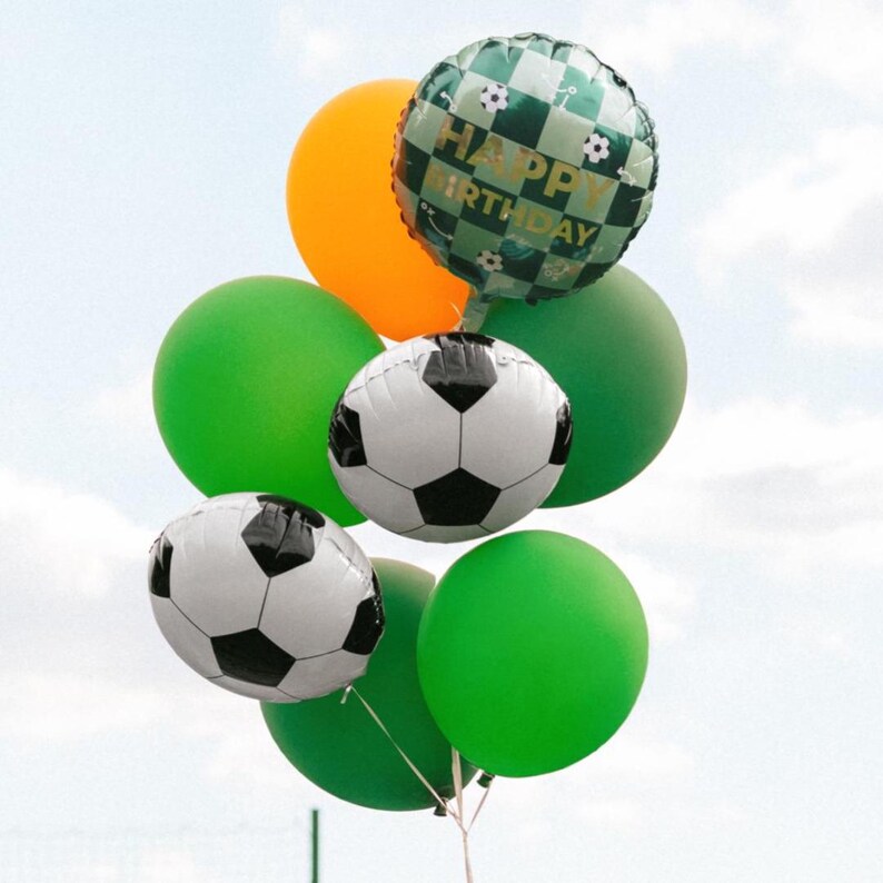 May include: A cluster of balloons in shades of green and orange, including two soccer ball-shaped balloons and a round balloon with "HAPPY BIRTHDAY" text. The balloons are tied together, floating against a cloudy sky.