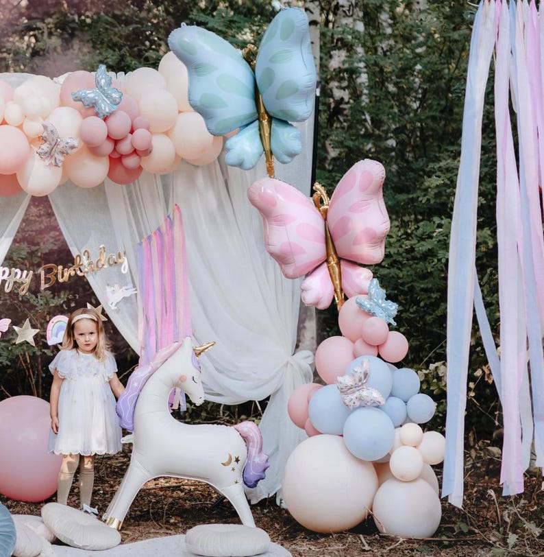 Pu&ograve; includere: Un animale a forma di unicorno in palloncino con corpo bianco, criniera e coda viola e zoccoli dorati. L'unicorno &egrave; circondato da palloncini rosa pastello, blu e bianchi, tra cui palloncini a forma di farfalla. Una bambina con un vestito bianco &egrave; in piedi accanto all'unicorno.