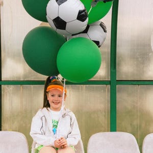 May include: A young person sits on a bench, holding a collection of balloons. The balloons include green, orange, and soccer ball-themed designs, with one reading "Happy Birthday". The person wears a white hoodie, green shorts, and a visor.