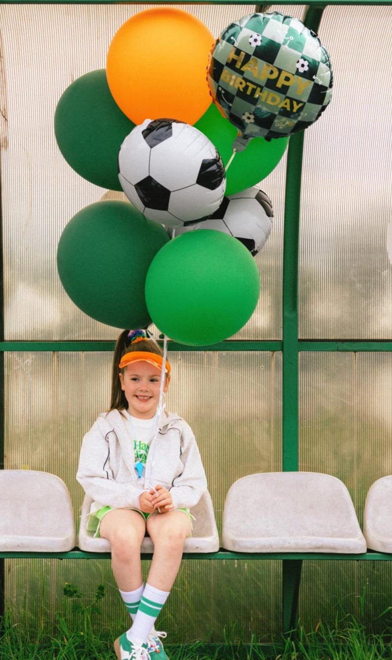 May include: A young person sits on a bench, holding a collection of balloons. The balloons include green, orange, and soccer ball-themed designs, with one reading "Happy Birthday". The person wears a white hoodie, green shorts, and a visor.