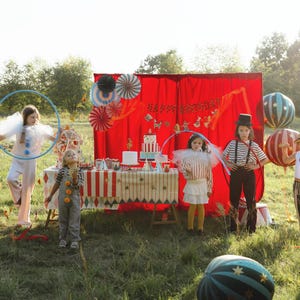 K&ouml;nnte beinhalten: Eine festliche Szene im Freien mit Kindern in Zirkus-Kost&uuml;men. Ein Tisch ist mit Kuchen und Dekorationen vor einem roten Hintergrund gedeckt. Gro&szlig;e, gestreifte Ballons und ein blauer Reifen tragen zur spielerischen Atmosph&auml;re bei.