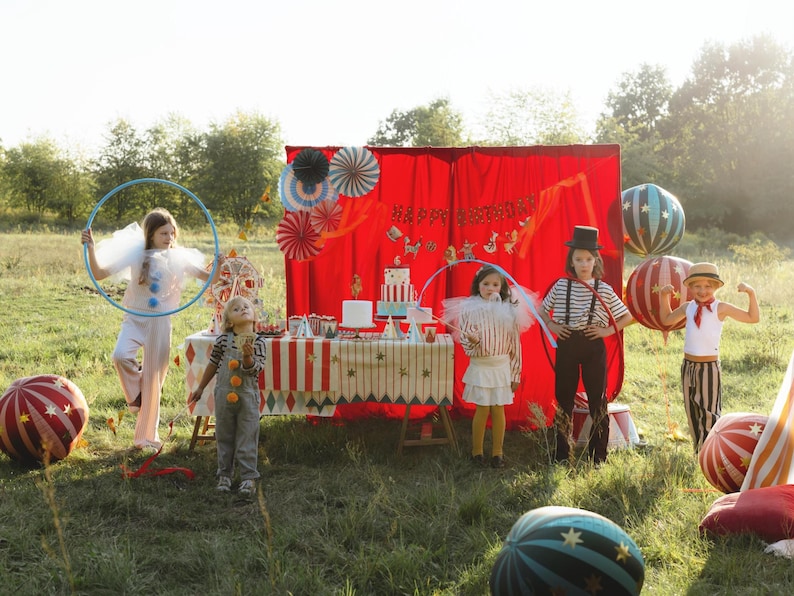 K&ouml;nnte beinhalten: Eine festliche Szene im Freien mit Kindern in Zirkus-Kost&uuml;men. Ein Tisch ist mit Kuchen und Dekorationen vor einem roten Hintergrund gedeckt. Gro&szlig;e, gestreifte Ballons und ein blauer Reifen tragen zur spielerischen Atmosph&auml;re bei.