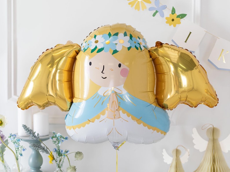 A gold and pastel-colored foil balloon shaped like a praying angel with golden wings, blonde hair, and a floral crown. The balloon is part of a soft, elegant party setup with pastel flowers, candles, and paper angel decorations in the background, perfect for a baptism, communion, or baby celebration.