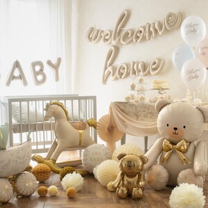 Op de afbeelding: Een babyshower-sc&egrave;ne met een wieg, een schommelpaard en ballonnen. De woorden "BABY" en "welcome home" worden weergegeven. Een grote teddybeerballon en een kleinere gouden teddybeerfiguur zijn te zien, samen met decoratieve papieren waaiers.