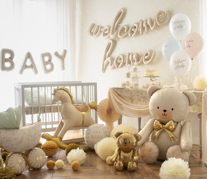 Op de afbeelding: Een babyshower-sc&egrave;ne met een wieg, een schommelpaard en ballonnen. De woorden "BABY" en "welcome home" worden weergegeven. Een grote teddybeerballon en een kleinere gouden teddybeerfiguur zijn te zien, samen met decoratieve papieren waaiers.