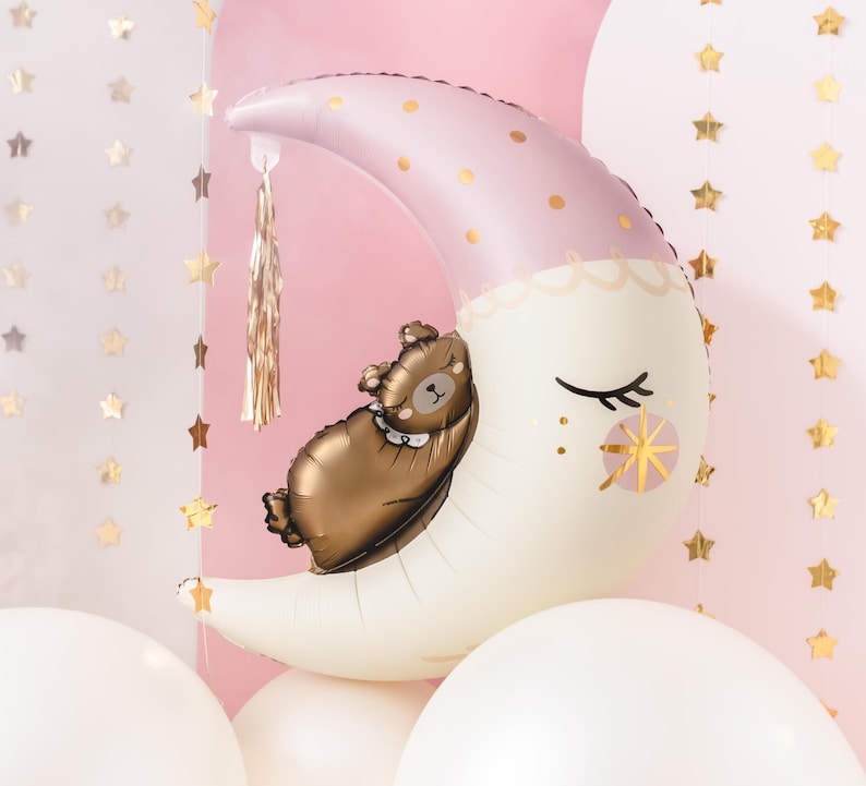 Pu&ograve; includere: Un palloncino a forma di luna crescente rosa e bianco con accenti di glitter dorati e un orso bruno che dorme sopra. Il palloncino &egrave; circondato da palloncini bianchi.