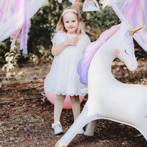 K&ouml;nnte beinhalten: Ein junges M&auml;dchen in einem wei&szlig;en Kleid steht neben einem gro&szlig;en aufblasbaren Einhorn. Das Einhorn ist wei&szlig; mit einer rosa-lila M&auml;hne und einem goldenen Horn. Ein rosa Ballon und ein dekoratives Baldachin befinden sich im Hintergrund.