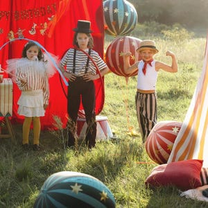 May include: Three children dressed in circus-themed costumes pose outdoors. One wears a top hat and striped shirt, another holds a hoop, and the third flexes their arm. Balloons and a red tent add to the festive atmosphere.