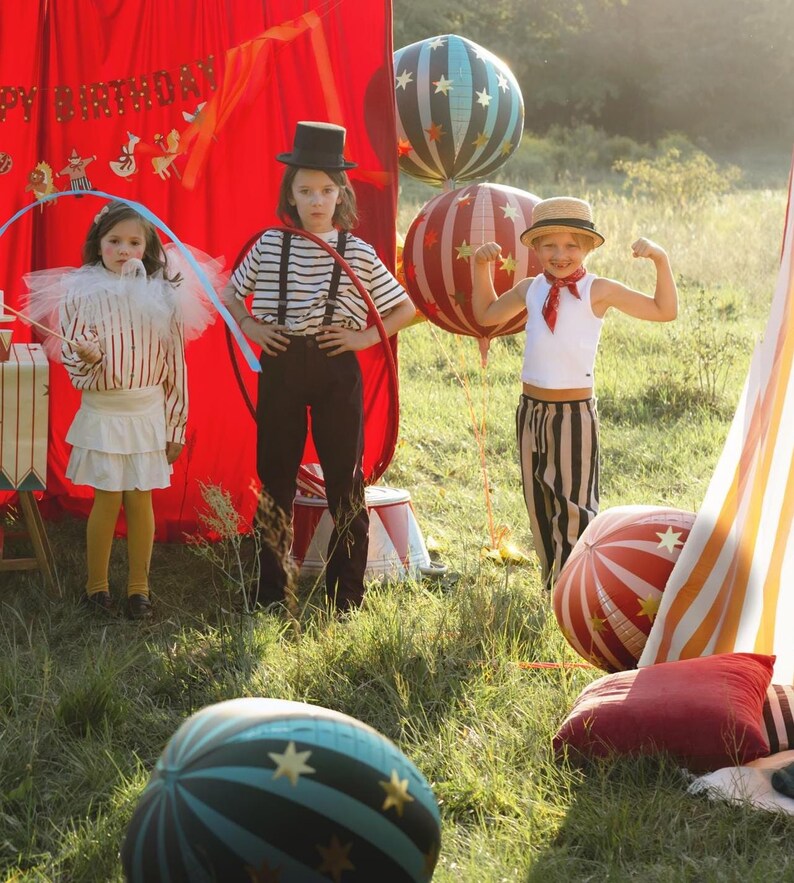 May include: Three children dressed in circus-themed costumes pose outdoors. One wears a top hat and striped shirt, another holds a hoop, and the third flexes their arm. Balloons and a red tent add to the festive atmosphere.