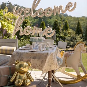 May include: A baby shower scene with a table set for a party. A gold teddy bear balloon and a rocking horse balloon are in the foreground. A balloon banner says "welcome home". The table is set with floral patterned dishes.