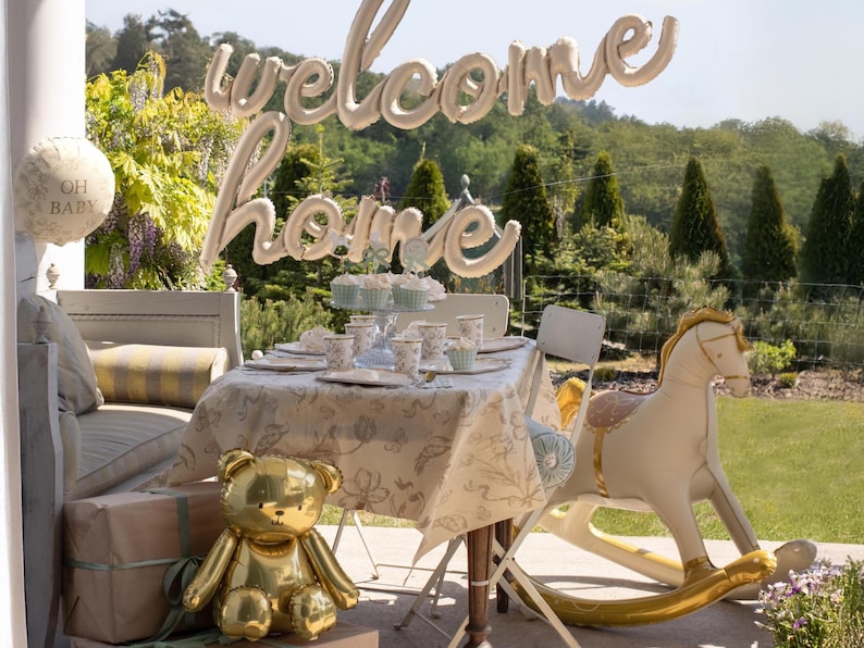 May include: A baby shower scene with a table set for a party. A gold teddy bear balloon and a rocking horse balloon are in the foreground. A balloon banner says "welcome home". The table is set with floral patterned dishes.