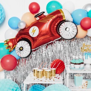 May include: Red, blue, and silver balloons with a red race car shaped balloon. The race car balloon has a lightning bolt and gold accents. There are also cupcakes with blue frosting and a cake with white frosting and a drip of blue icing. The text on the cake says "Happy Birthday".