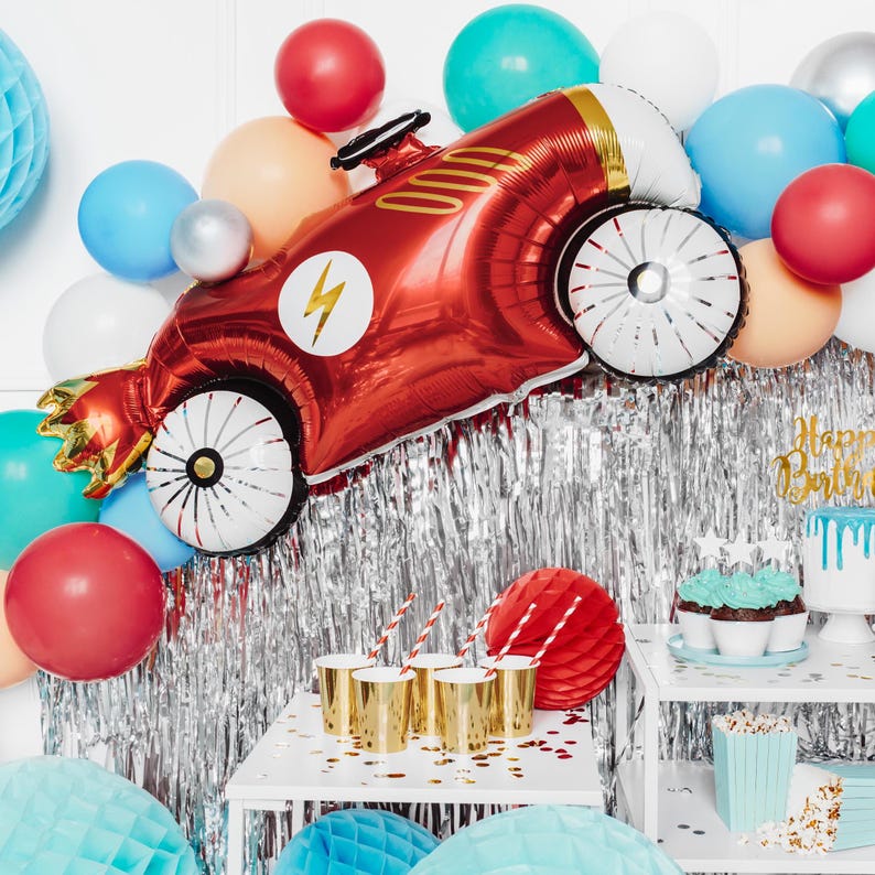 May include: Red, blue, and silver balloons with a red race car shaped balloon. The race car balloon has a lightning bolt and gold accents. There are also cupcakes with blue frosting and a cake with white frosting and a drip of blue icing. The text on the cake says "Happy Birthday".