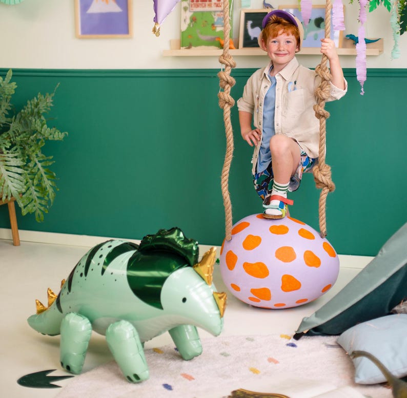 Puede incluir: Un dinosaurio inflable verde y dorado con un huevo inflable de lunares morados y naranjas. Un ni&ntilde;o peque&ntilde;o con una camisa azul y una chaqueta beige est&aacute; de pie sobre el huevo.