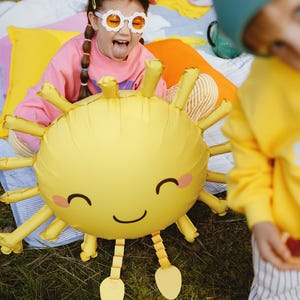 Children at an outdoor party playing with a smiling sun-shaped balloon. A girl in colorful clothes and fun sunglasses poses happily behind the balloon, while a boy in a beanie and bright outfit appears in the foreground. Bright pillows and picnic setup in the background create a cheerful, sunny atmosphere.