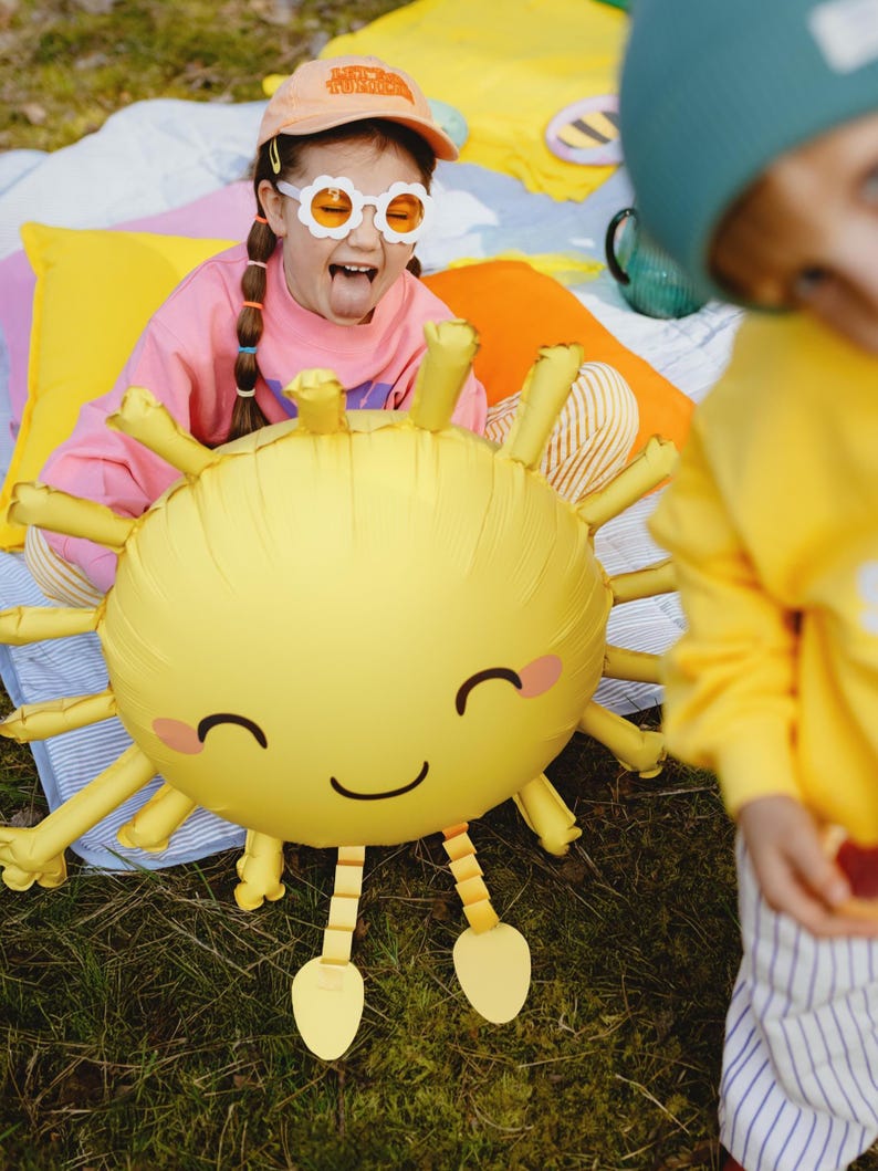 Children at an outdoor party playing with a smiling sun-shaped balloon. A girl in colorful clothes and fun sunglasses poses happily behind the balloon, while a boy in a beanie and bright outfit appears in the foreground. Bright pillows and picnic setup in the background create a cheerful, sunny atmosphere.
