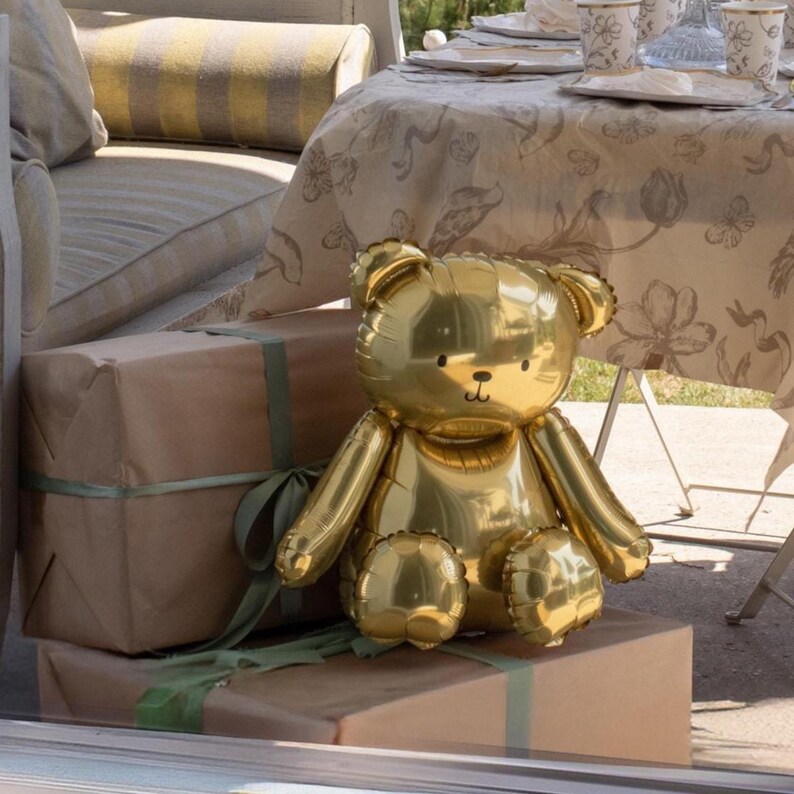 May include: A shiny gold teddy bear balloon sits atop a stack of brown wrapped gifts tied with green ribbon. The bear has a smiling face and is positioned in front of a table set for a party.