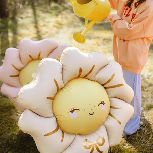 Puede incluir: Una ni&ntilde;a con un sombrero azul y una sudadera naranja riega dos grandes globos de flores inflables con una regadera amarilla. Los globos tienen forma de girasoles con caras sonrientes y detalles dorados.