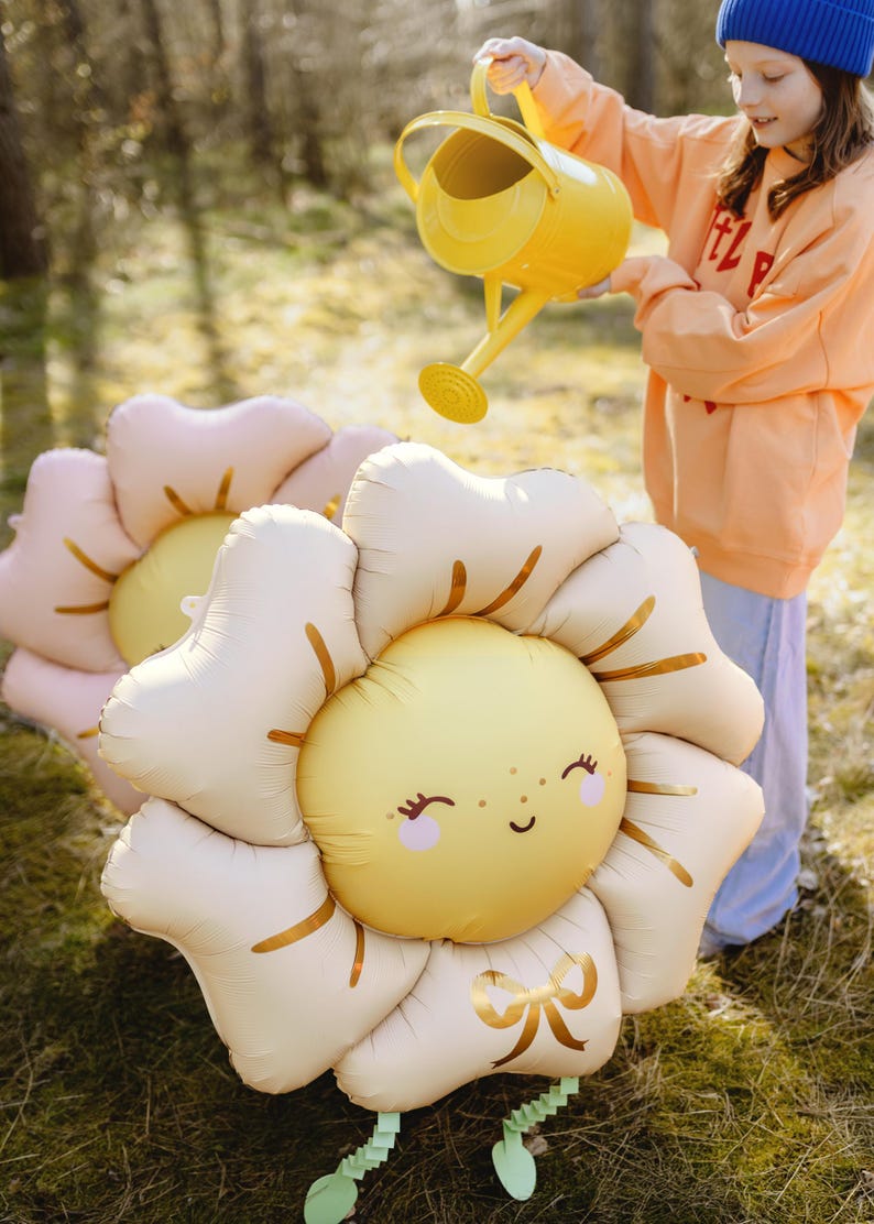 Puede incluir: Una ni&ntilde;a con un sombrero azul y una sudadera naranja riega dos grandes globos de flores inflables con una regadera amarilla. Los globos tienen forma de girasoles con caras sonrientes y detalles dorados.