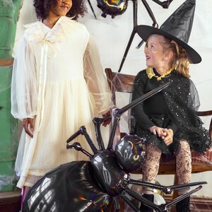 Op de afbeelding: Twee kinderen gekleed in Halloween-kostuums. E&eacute;n kind draagt een witte jurk met een bloemenkrans en het andere draagt een zwart heksenkostuum met een zwarte hoed. Beide kinderen kijken naar een grote zwarte spinballon.