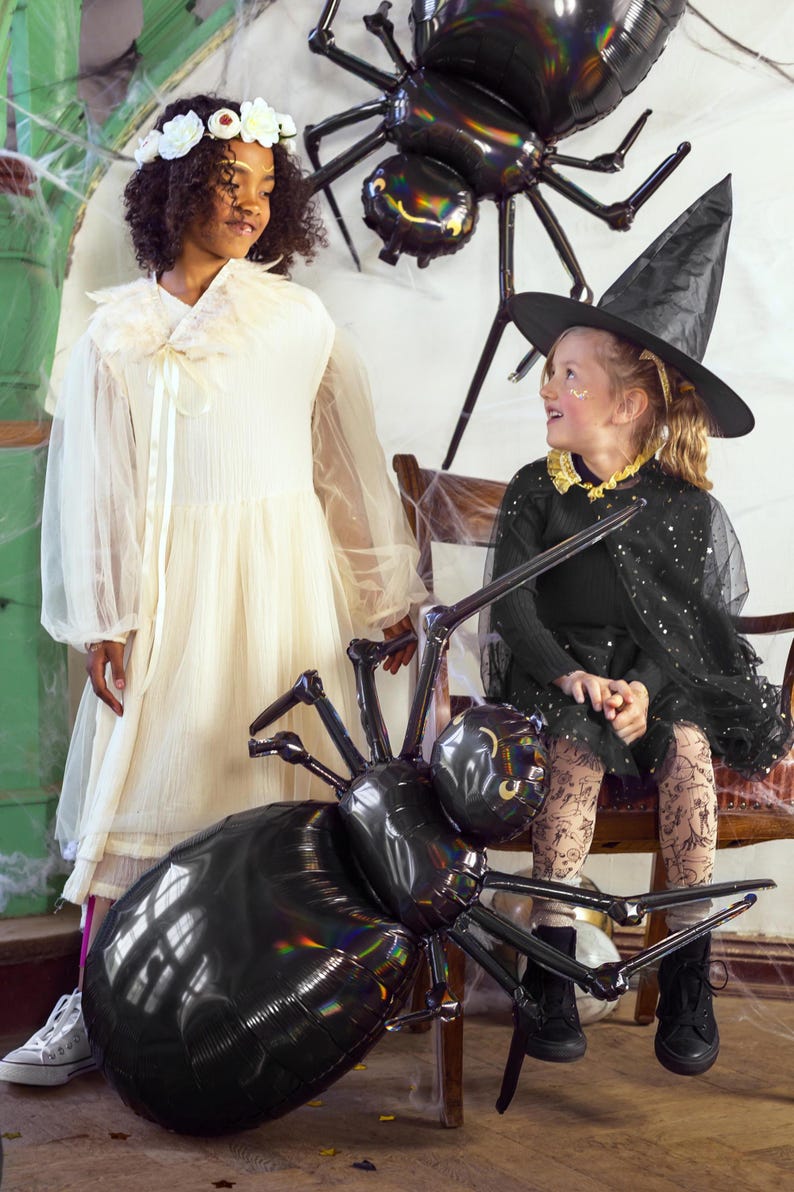 Op de afbeelding: Twee kinderen gekleed in Halloween-kostuums. E&eacute;n kind draagt een witte jurk met een bloemenkrans en het andere draagt een zwart heksenkostuum met een zwarte hoed. Beide kinderen kijken naar een grote zwarte spinballon.