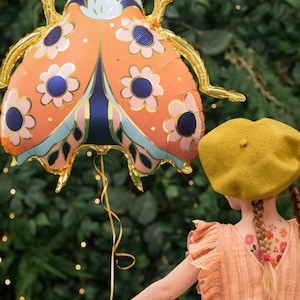 May include: A young person holds a large orange and gold ladybug balloon with a floral pattern. The balloon is held by a gold ribbon. The person is wearing a yellow beret and a striped dress.