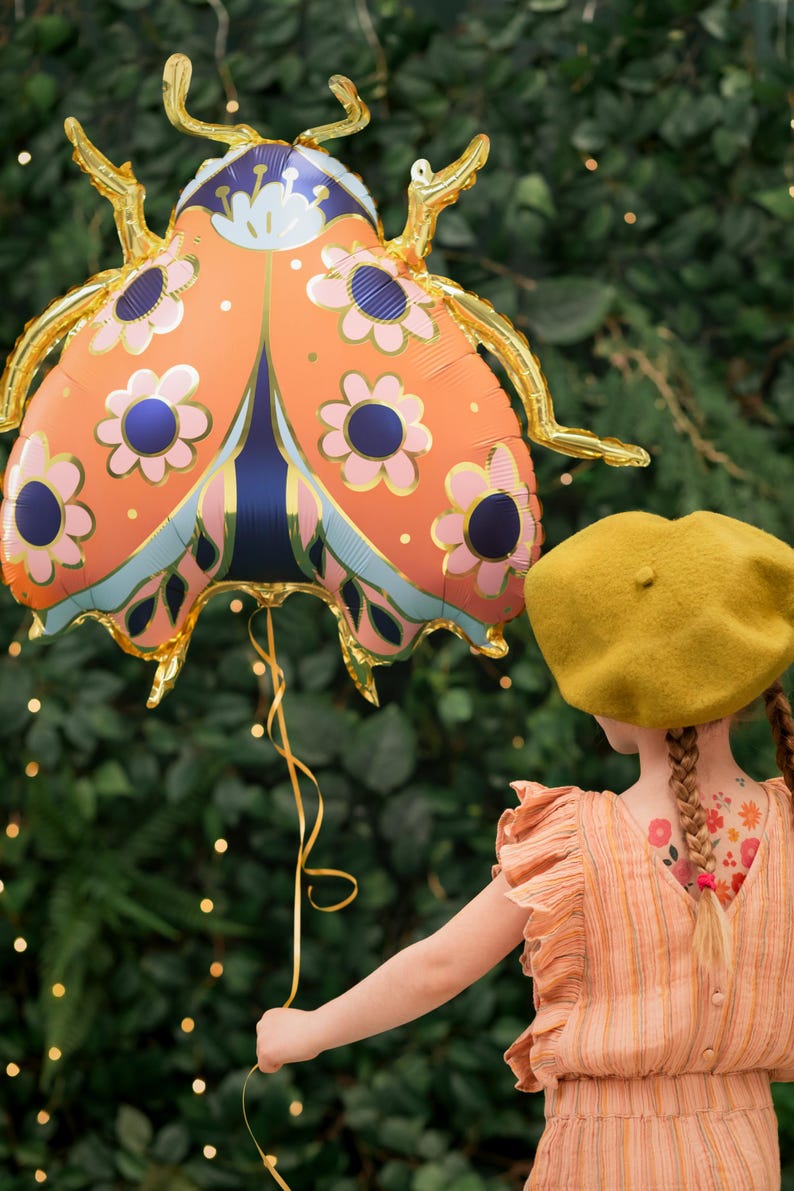 May include: A young person holds a large orange and gold ladybug balloon with a floral pattern. The balloon is held by a gold ribbon. The person is wearing a yellow beret and a striped dress.