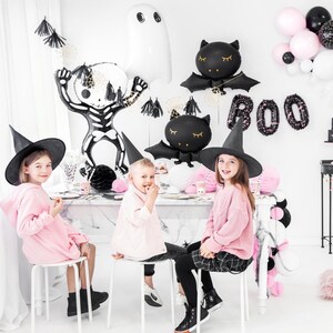 Puede incluir: Tres ni&ntilde;os con sombreros de bruja y ropa rosa est&aacute;n sentados en una mesa decorada para una fiesta de Halloween. La mesa est&aacute; decorada con globos negros y blancos, incluyendo un esqueleto, un fantasma y murci&eacute;lagos. La mesa tambi&eacute;n est&aacute; decorada con serpentinas negras y blancas y una pancarta negra y blanca que dice "BOO".
