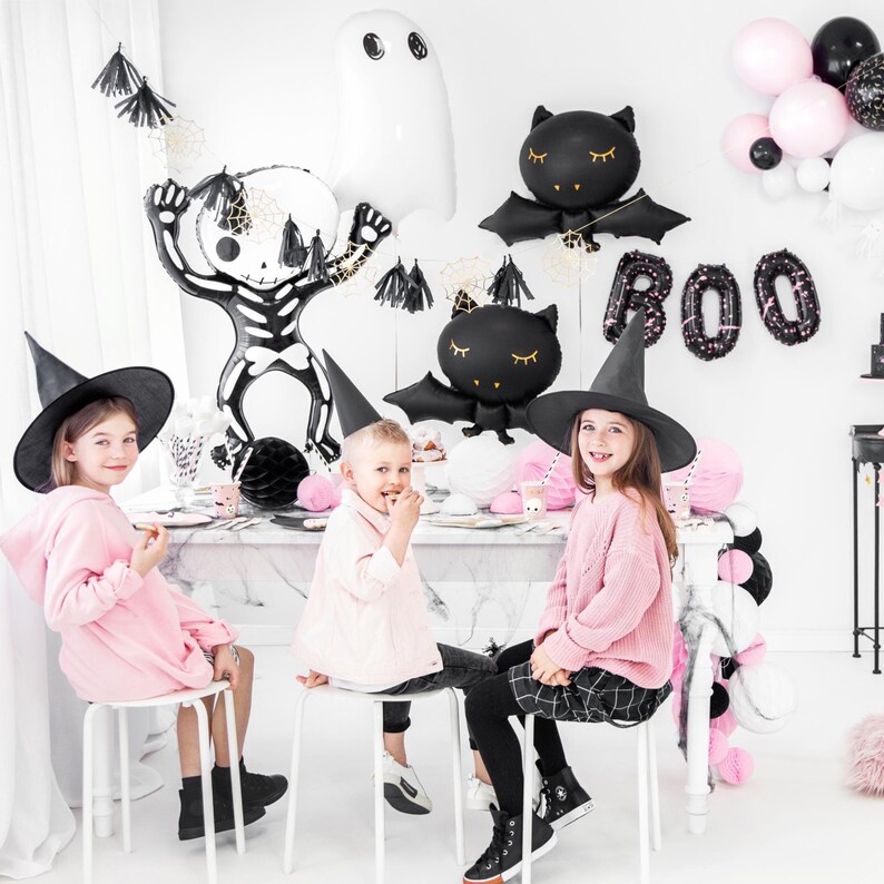 Puede incluir: Tres ni&ntilde;os con sombreros de bruja y ropa rosa est&aacute;n sentados en una mesa decorada para una fiesta de Halloween. La mesa est&aacute; decorada con globos negros y blancos, incluyendo un esqueleto, un fantasma y murci&eacute;lagos. La mesa tambi&eacute;n est&aacute; decorada con serpentinas negras y blancas y una pancarta negra y blanca que dice "BOO".