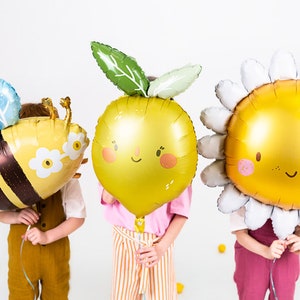 K&ouml;nnte beinhalten: Drei Kinder halten bunte Folienballons in Form einer Biene, einer Zitrone und einer G&auml;nsebl&uuml;mchen. Der Bienenballon ist gelb und schwarz mit blauen Fl&uuml;geln. Der Zitronenballon ist gelb mit einem gr&uuml;nen Blatt und einem l&auml;chelnden Gesicht. Der G&auml;nsebl&uuml;mchenballon ist wei&szlig; mit einer gelben Mitte und einem l&auml;chelnden Gesicht.