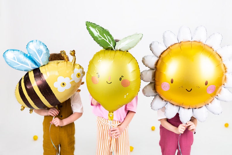 K&ouml;nnte beinhalten: Drei Kinder halten bunte Folienballons in Form einer Biene, einer Zitrone und einer G&auml;nsebl&uuml;mchen. Der Bienenballon ist gelb und schwarz mit blauen Fl&uuml;geln. Der Zitronenballon ist gelb mit einem gr&uuml;nen Blatt und einem l&auml;chelnden Gesicht. Der G&auml;nsebl&uuml;mchenballon ist wei&szlig; mit einer gelben Mitte und einem l&auml;chelnden Gesicht.