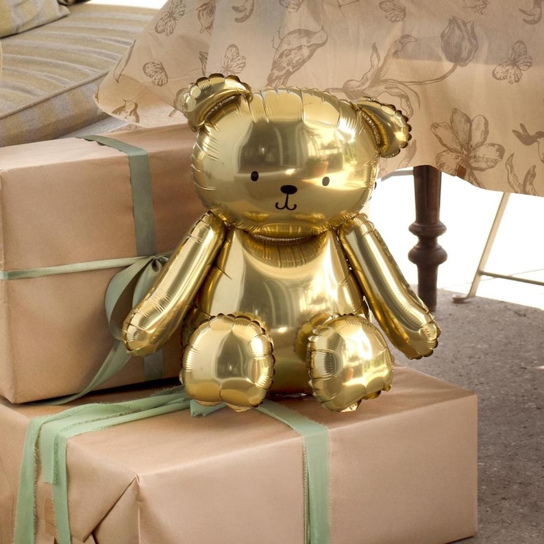 May include: A shiny gold teddy bear balloon sits atop wrapped gift boxes with green ribbon. The bear has a classic design with rounded ears, arms, and legs. The background includes a floral patterned tablecloth and a wooden table leg.