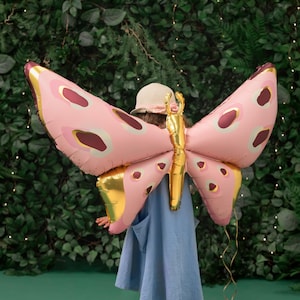 May include: A child wearing a blue dress and a straw hat holds a large pink and gold butterfly balloon behind their back. The balloon has a gold body and pink wings with brown and white spots.
