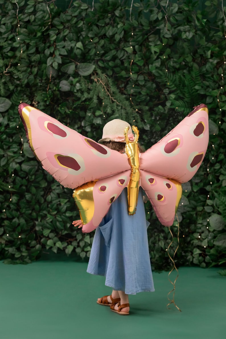 May include: A child wearing a blue dress and a straw hat holds a large pink and gold butterfly balloon behind their back. The balloon has a gold body and pink wings with brown and white spots.