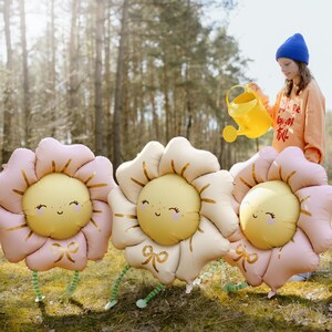 Puede incluir: Tres grandes globos de flores inflables con p&eacute;talos rosas y beige y caras sonrientes amarillas. Los globos tienen lazos dorados y tallos verdes. Una persona est&aacute; regando los globos con una regadera amarilla.