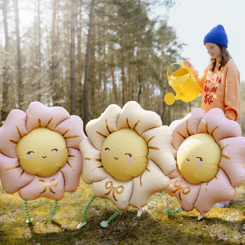 Puede incluir: Tres grandes globos de flores inflables con p&eacute;talos rosas y beige y caras sonrientes amarillas. Los globos tienen lazos dorados y tallos verdes. Una persona est&aacute; regando los globos con una regadera amarilla.