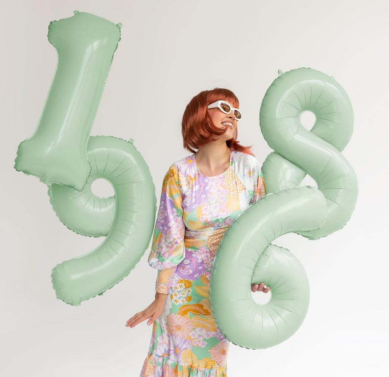 Pu&ograve; includere: Una persona con un vestito floreale e occhiali da sole tiene in mano due grandi palloncini numerati in lamina verde chiaro che compongono la scritta "18".