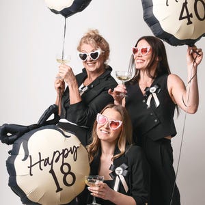 May include: Three women wearing black outfits and sunglasses pose with gold and black balloons that say "Happy 18", "Happy 40", and "Happy 60".