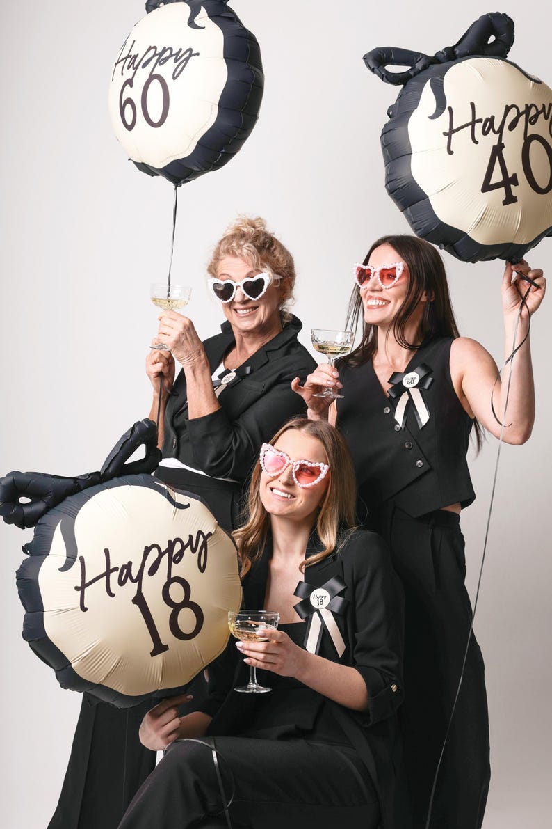 May include: Three women wearing black outfits and sunglasses pose with gold and black balloons that say "Happy 18", "Happy 40", and "Happy 60".